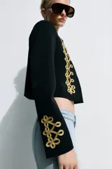 Blazer crop color negro, con cuello subido, manga larga y cierre frontal con botones ocultos. Presenta detalles de alamares bordados a contraste en color dorado en el frente y en los puños.