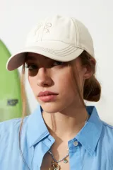 Gorra de gabardina color crudo con pespuntes en contraste y moño bordado en el frente.