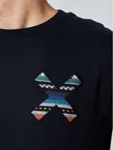 Remera blanca de algodón orgánico con cuello redondo y manga corta. Presenta un estampado en el pecho con una cruz formada por dos tablas de surf en tonos rojo, azul y negro.
