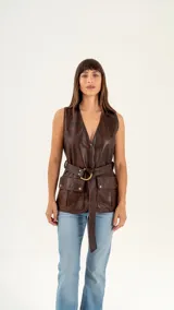 Chaleco largo marrón de cuero vacuno gastado, con cierre frontal con cremallera, cinturón con hebilla metálica y bolsillos amplios con solapa.
