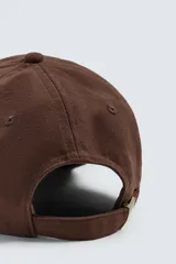 Gorra de seis paneles confeccionada en tejido de algodón, color marrón, con bordado frontal en color crema que incluye texto. Cuenta con ajuste en la parte posterior.