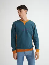 Buzo azul con cuello redondo, mangas largas y puños y bajo acanalados. Presenta bloque de color naranja en hombros y laterales, y estampado frontal con texto en color borgoña.