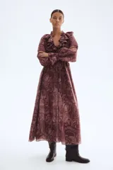 Vestido largo de manga globo, color marrón oscuro, con estampado paisley en tonos claros. Presenta cuello alto con volados y escote en V con lazos ajustables, y puños con volados.