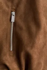 Campera de gamuza color marrón con acabado cepillado suave, cuello clásico, hombros caídos y mangas largas. Cuenta con cierre frontal oculto bajo solapa con botones a presión, bolsillos laterales con cierre y diseño abullonado con elástico interior en el bajo.
