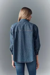 Camisa de denim azul con apliques metálicos plateados y bordado de piedras rojas y negras en los bolsillos delanteros. Tiene calce amplio, cuello camisero y cierre con botones a presión.