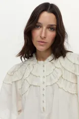 Blusa blanca con cuello mao, detalles de mini volados y bordados en el pecho y las mangas, con cierre frontal de botones y mangas largas abullonadas.