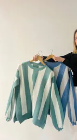 Sweater de punto con diseño de rayas verticales en tonos azul y gris, con sutil acabado brillante. Presenta cuello redondo, mangas largas con puños ajustados y corte amplio con aberturas laterales en el ruedo.