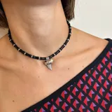 Gargantilla de cuentas cilíndricas negras y detalles plateados, con dije colgante de diente de tiburón plateado.