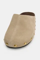 Zapato destalonado tipo mule de serraje en color beige, con punta redonda, suela plana y detalle de tachas metálicas doradas alrededor de la plataforma.