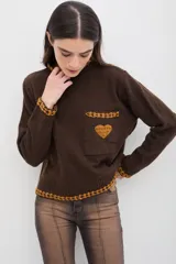 Sweater de punto color marrón con cuello alto, manga larga y calce relajado. Presenta detalles bordados en color mostaza en el cuello, puños, ruedo y bolsillo frontal, además de un aplique en forma de corazón en el bolsillo.