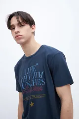 Remera de algodón color azul marino con cuello redondo y mangas cortas. Presenta un estampado gráfico central con texto en tonos azul claro, rojo y amarillo, incluyendo una pequeña estrella.