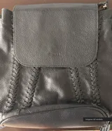 Cartera negra de cuero convertible en mochila, con tapa superior, cierre metálico, terminaciones trenzadas, forro de gabardina de algodón, bolsillos internos y externos con cierre.