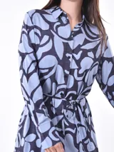 Vestido camisero midi de manga larga, con cuello y botones frontales, ajustado a la cintura con un lazo. Presenta un estampado abstracto en tonos azul claro y azul oscuro/negro.