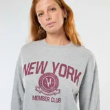 Buzo de cuello redondo en color gris melange, con mangas largas y corte holgado. Presenta un estampado frontal con la inscripción 'NEW YORK' y un logo circular estilo universitario con el texto 'MEMBER CLUB' en color bordó.