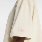 Remera corta de manga corta, color beige claro, con cuello redondo. Presenta un estampado frontal colorido que muestra un par de zapatillas con calcetines a rayas de colores vibrantes (amarillo, fucsia, azul) y el texto "new balance" debajo.