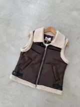 Chaleco de cuero sintético en color marrón con interior y bordes de corderito sintético color crema. Presenta cierre frontal, bolsillos laterales y detalles de correas ajustables con hebillas en la parte inferior.