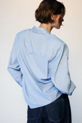 Camisa de manga larga con cuello solapa y hombreras. Presenta un diseño de rayas verticales en tonos azul y blanco, con cierre frontal de botones y puños anchos.