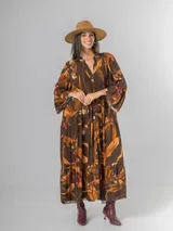 Vestido largo de manga larga, corte oversized y cuello camisero con abertura en V y botones. Presenta un estampado botánico en tonos tierra sobre fondo marrón oscuro, con volados en el ruedo.