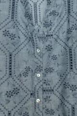 Camisa azul con cuello de solapa, escote en pico y mangas largas con puños abotonados. Presenta un diseño con bordados perforados y patrones geométricos y florales. Cierre frontal con botones.