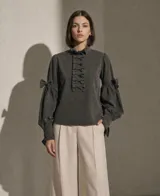 Blusa de color verde oscuro con cuello alto con volados, detalle de lazos frontales y mangas largas abullonadas con lazos decorativos en los codos y puños ajustados.