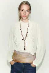 Blusa de bambula color blanco con cuello subido y manga larga. Presenta detalles de volantes y puntillas a tono en el frente, con cierre posterior mediante abertura y botón.