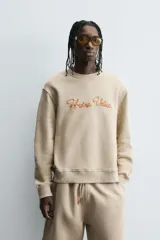 Sudadera de algodón color beige claro, corte relajado, cuello redondo y manga larga. Presenta bordado en relieve en el frente con texto en cursiva de color cobre.