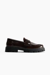 Mocasines tipo penny de color marrón oscuro, confeccionados en material sintético con acabado brillante. Presentan flecos decorativos en el empeine, una banda transversal con detalle metálico y una suela gruesa dentada de color negro.