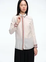 Camisa de paño color crudo con detalles bordados en tonos rojizos y marrones en el cuello, la botonera frontal y los puños. Presenta un corte recto y cómodo con mangas largas.