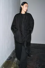 Chaqueta oversize de nylon en color negro, con cuello redondo subido, manga larga y cierre frontal con botones a presión ocultos. Presenta bolsillos delanteros y forro interior a tono.