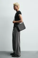 Bandolera de diseño rectangular en color negro con acabado texturizado. Cuenta con una solapa frontal con detalle metálico, cierre imantado y asa de hombro ajustable. El interior incluye un bolsillo plano y otro con cierre, además de un bolsillo plano adicional en la parte frontal.