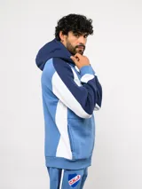 Campera deportiva Umbro del Club Nacional de Football, con cierre frontal, capucha ajustable con cordones y diseño en bloques de color azul, celeste y blanco. Presenta el logo de la marca y el escudo del club bordados en el pecho.