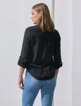 Blusa blanca de gasa marca Zac & Rachel, con cuello a la base, escote en V, abertura central con botones, bolsillo aplicado en el pecho y mangas largas regulables con presilla y botón. Tiene terminación opcional de nudo en el ruedo.