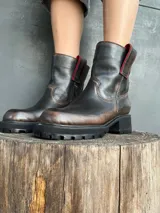 Bota de cuero con efecto desgastado en tonos marrones y negros, con suela track robusta y cierre lateral. Presenta detalles en color rojo en la parte superior de la caña.