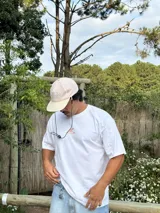 Gorro de visera curva con diseño a rayas verticales en tonos rosado pálido y blanco. La corona tiene un bordado en azul claro con texto. La visera es de color beige claro. Incluye un cordón negro ajustable para colgar del cuello.