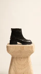Bota corta de cuero negro con diseño de paneles que combina cuero liso y gamuza. Presenta puntera cuadrada, cierre trasero y suela con plataforma y taco bajo geométrico.