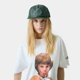 Gorra verde oscura tipo trucker con frente de cuerina, visera plana y paneles traseros de malla. Cuenta con escudo bordado en la parte frontal y cierre snapback ajustable.