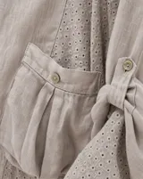 Blusa beige de lino de corte holgado y silueta oversize, con cuello redondo, cierre frontal de botones, bolsillos aplicados grandes y mangas tres cuartos con detalles de perforaciones bordadas en las mangas y el ruedo.