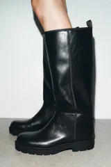 Bota de caña alta estilo plano, confeccionada en material sintético negro con acabado liso. Presenta detalle de pespuntes verticales en el cuerpo, punta redonda, suela track dentada y tirador trasero para facilitar el calce.