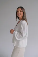 Blusa blanca de corte holgado con cuello en V, cierre frontal de botones y mangas largas abullonadas con puños elásticos. Presenta detalles bordados sutiles en la parte frontal y en las mangas.