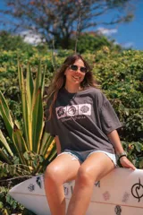 Remerón cropped gris oscuro con estampado frontal de tres recuadros en rosa pastel. Los recuadros contienen un hibisco, una ola y una concha marina, con el texto "Glassy Waves" debajo.