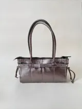 Cartera de cuero de tamaño medio con estructura rígida. Presenta una tira decorativa con tachas y hebillas metálicas en los extremos, además de lazos laterales. Cuenta con doble asa para llevar al hombro y cierre superior.