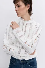 Camisa de algodón color blanco con detalles de encaje broderie y alforzas verticales. Presenta cuello alto con volados, cierre frontal de botones y mangas largas con puños acampanados.