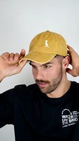 Gorra tipo baseball color mostaza con efecto desgastado, confeccionada en algodón. Presenta un bordado pequeño en color blanco en el panel frontal que representa un pájaro con sombrero sobre una rama.