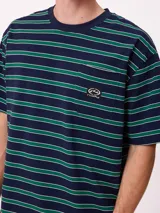 Remera de corte holgado con diseño de rayas horizontales en tonos beige, marrón y azul claro. Presenta cuello redondo, mangas cortas y un bolsillo frontal en el pecho con el logo de la marca bordado.