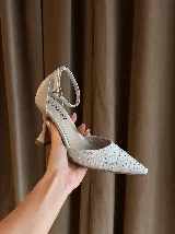 Zapato de tacón con punta fina, confeccionado en tela con aplicación de strass en la capellada y detalles en cuero en el talón, taco y pulsera. Cuenta con cierre de hebilla al tobillo y taco de altura media.