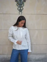 Camisa de lino color beige, con cuello bobo y detalles de encaje bordado en el cuello, pechera y puños. Cuenta con mangas largas levemente abuchonadas y cierre frontal con botones nacarados.