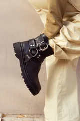 Bota corta negra de cuero sintético con suela track, caña baja y correas con hebillas metálicas.