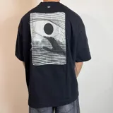 Remera de corte oversize confeccionada en algodón pesado, color negro. Presenta un parche rectangular cosido en la espalda con un diseño gráfico artístico en blanco y negro que muestra una mano alcanzando un círculo oscuro sobre un fondo de líneas horizontales.
