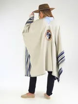 Poncho grueso color beige con detalles de rayas azules en los laterales y flecos en los bordes. Presenta el escudo bordado en la espalda y diseño liso en la parte frontal con escote en V.
