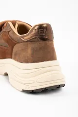 Zapatilla urbana de cuero lavado en tonos marrones, con paneles de gamuza y detalles metalizados en el talón. Presenta cordones al tono y una suela de plataforma de goma en color crema.